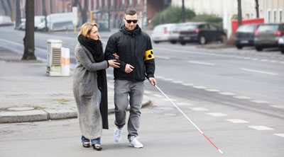 Visually impaired man walking across a road with a white stick and support from a woman